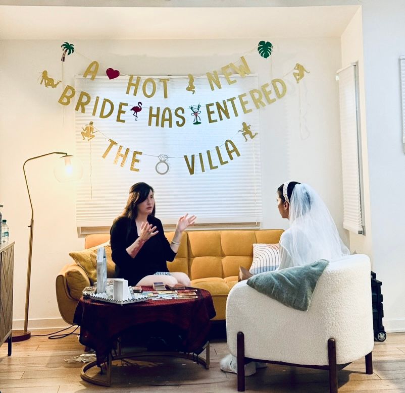 Bride-to-be wearing a veil sits in a modern living room for a bridal consultation with a friend on a mustard sofa beneath a gold “A hot new bride has entered the villa” banner, coffee table with wedding magazines and decor.