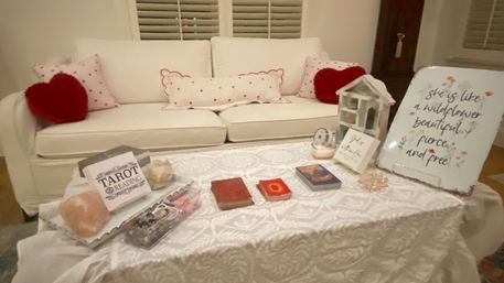 Cozy living room tarot-reading setup on a lace-covered coffee table in front of a white sofa with heart pillows — tarot decks, crystals, and a floral wildflower decor sign.
