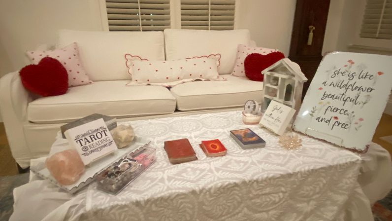 Cozy living room tarot-reading setup on a lace-covered coffee table in front of a white sofa with heart pillows — tarot decks, crystals, and a floral wildflower decor sign.