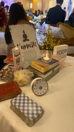 Tarot reading table at an indoor event: distressed “Namaste” sign, tarot card decks, crystal cluster, candle in a glass holder, timer, yellow flowers and a small “Tarot Reading” sign on a white tablecloth, guests seated in the background.