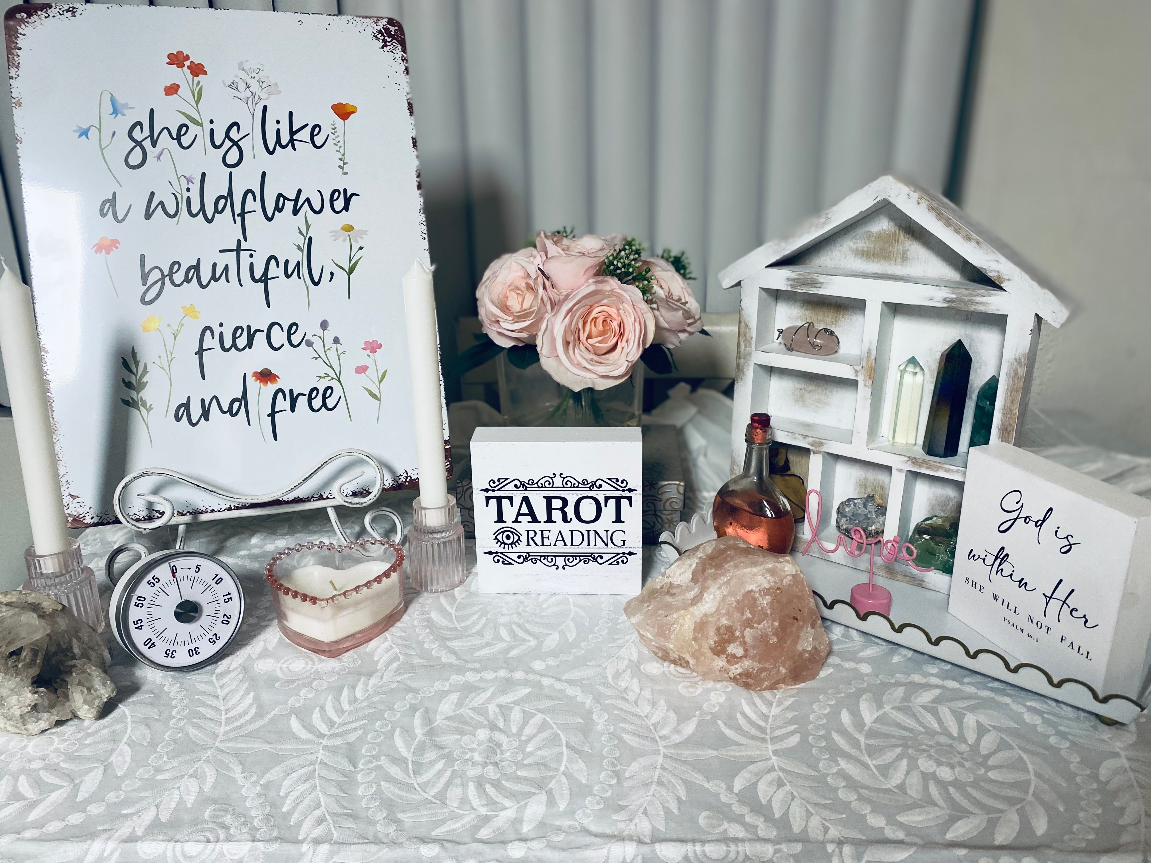 Boho tabletop spiritual display: white floral sign reading “she is like a wildflower,” pink rose bouquet, a block labeled TAROT READING, candles, crystal clusters and a salt lamp on a white patterned tablecloth.
