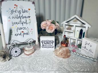 Boho tabletop spiritual display: white floral sign reading “she is like a wildflower,” pink rose bouquet, a block labeled TAROT READING, candles, crystal clusters and a salt lamp on a white patterned tablecloth.