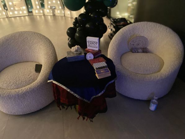 Cozy tarot reading nook at an indoor event: two cream boucle chairs flanking a small round table draped in blue and red cloth with tarot decks, crystal and a "Tarot Reading" sign, black balloon column and ambient lights.
