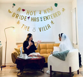 Cozy bachelorette party in a living room: bride in a veil on a white armchair talks with a friend on a mustard sofa under a gold “A HOT NEW BRIDE HAS ENTERED THE VILLA” banner, coffee table with party decorations and a floor lamp.