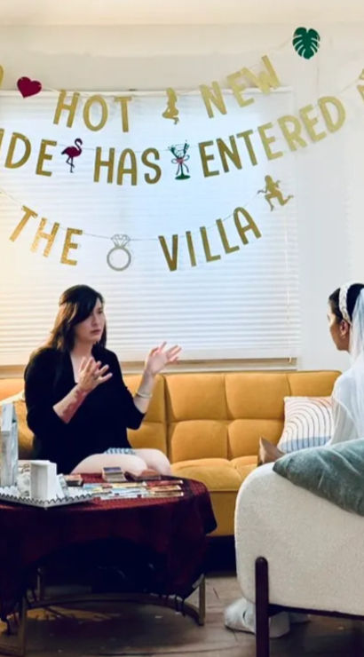 Cozy bachelorette party in a living room: bride in a veil on a white armchair talks with a friend on a mustard sofa under a gold “A HOT NEW BRIDE HAS ENTERED THE VILLA” banner, coffee table with party decorations and a floor lamp.