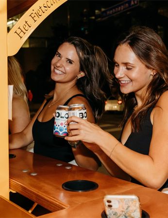 Two smiling women clinking colorful canned drinks over a wooden outdoor bar with cup holders on a nighttime city street