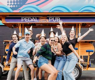 Group of friends in silver party hats laughing and celebrating in front of a bright orange pedal-powered party bike with a colorful mural backdrop on an urban street.