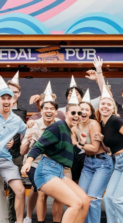 Group of friends in silver party hats laughing and celebrating in front of a bright orange pedal-powered party bike with a colorful mural backdrop on an urban street.