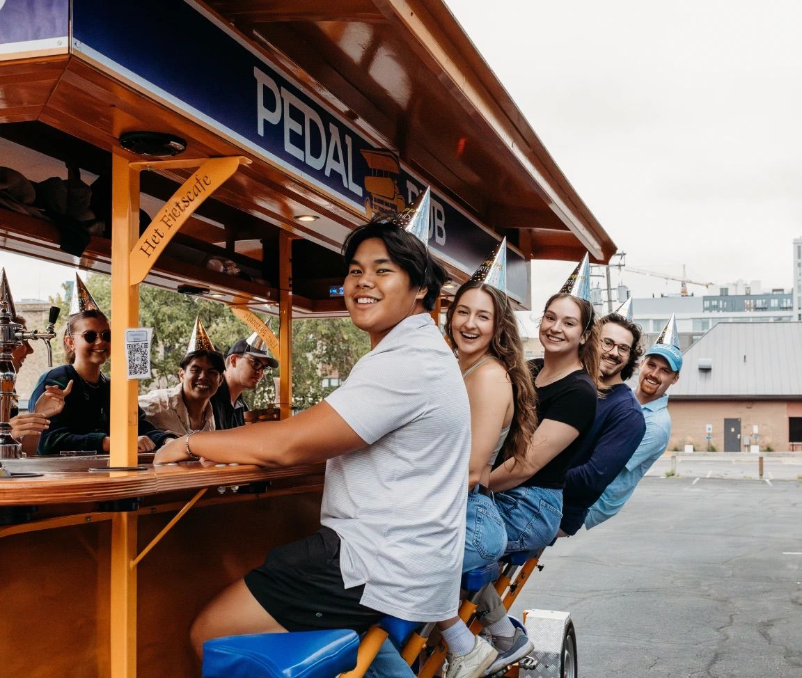 Smiling group of friends in party hats riding a pedal-powered bar bike outdoors in a downtown parking lot, enjoying a daytime celebration.
