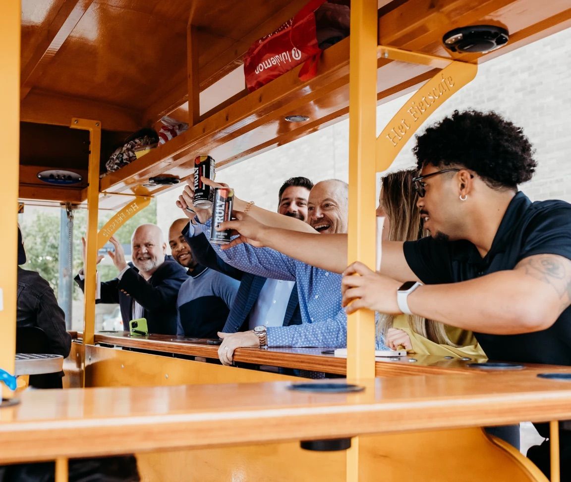 Friends cheering with canned drinks aboard a bright yellow pedal pub mobile bar, laughing during an outdoor urban pub crawl