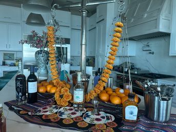 Modern white kitchen island with bright marigold garlands draped on a chrome hanging scale, bowls of oranges, liquor bottles and bar tools on a colorful beaded runner, large decorative vase in background.