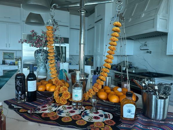 Modern white kitchen island with bright marigold garlands draped on a chrome hanging scale, bowls of oranges, liquor bottles and bar tools on a colorful beaded runner, large decorative vase in background.