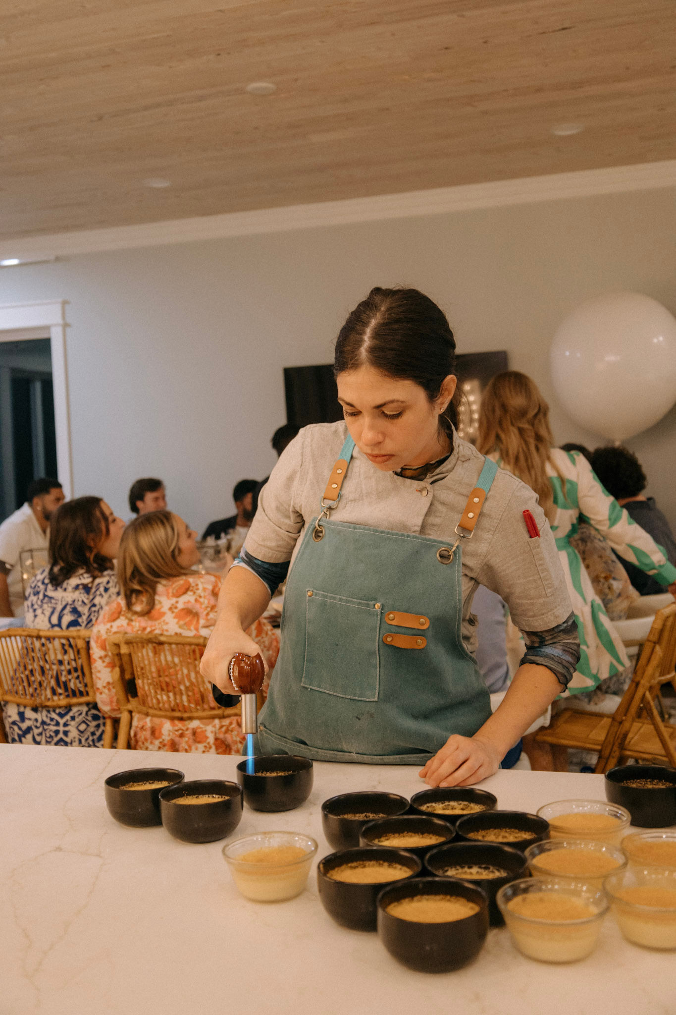 Chef using a culinary torch to caramelize crème brûlée in black and glass bowls on a marble kitchen island, preparing desserts for a cozy home dinner party with seated guests in the background.