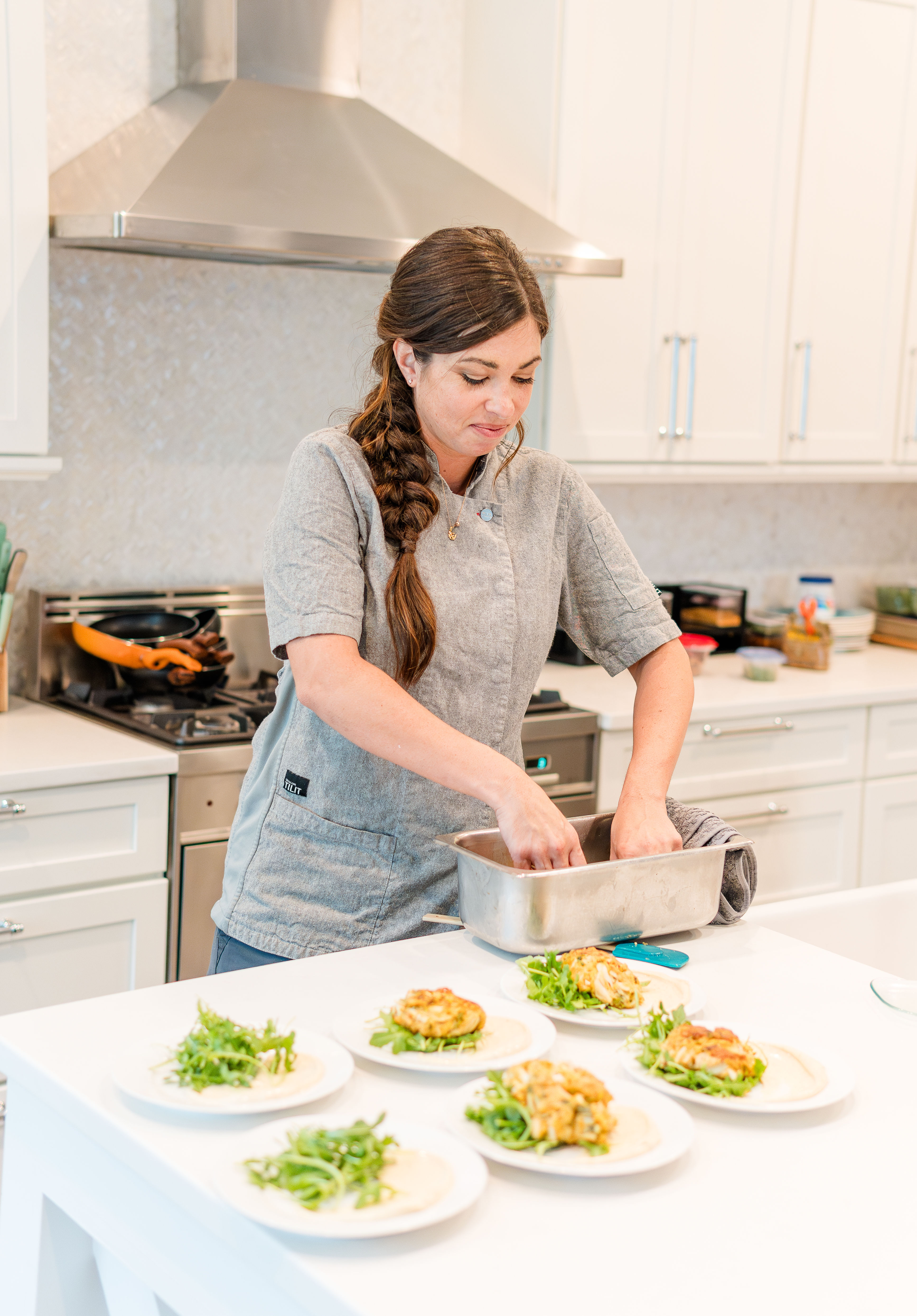 Home chef in a gray jacket with a braid plating golden fritters and fresh greens on white plates at a bright modern kitchen island under a stainless range hood.