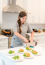 Home chef in a gray jacket with a braid plating golden fritters and fresh greens on white plates at a bright modern kitchen island under a stainless range hood.