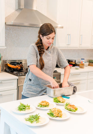 Home chef in a gray jacket with a braid plating golden fritters and fresh greens on white plates at a bright modern kitchen island under a stainless range hood.