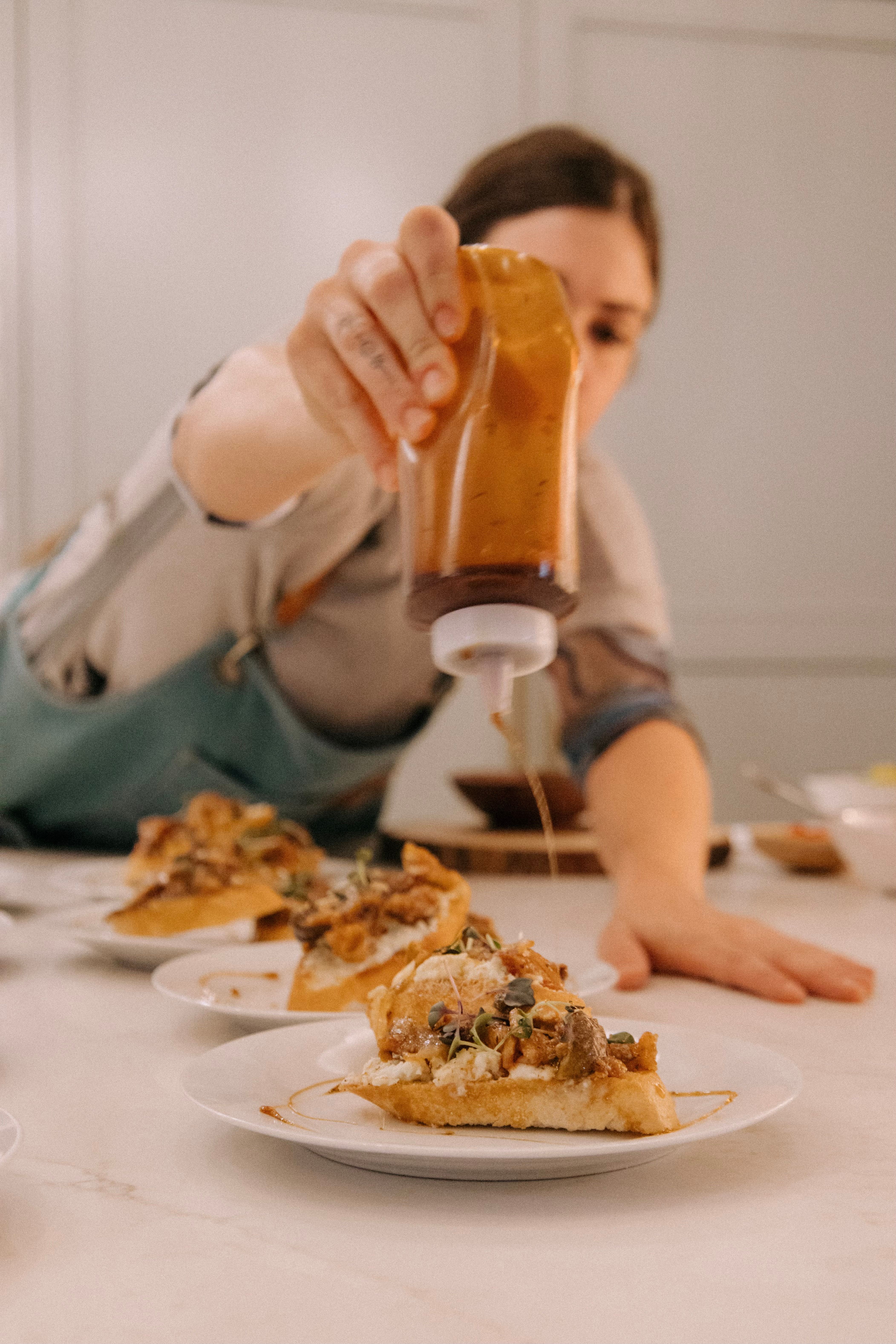 Apprent cook in an apron drizzling a caramel-colored sauce from a squeeze bottle onto toasted crostini topped with cheese, nuts and microgreens, plated on white dishes in a bright kitchen.