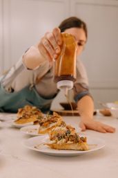 Apprent cook in an apron drizzling a caramel-colored sauce from a squeeze bottle onto toasted crostini topped with cheese, nuts and microgreens, plated on white dishes in a bright kitchen.