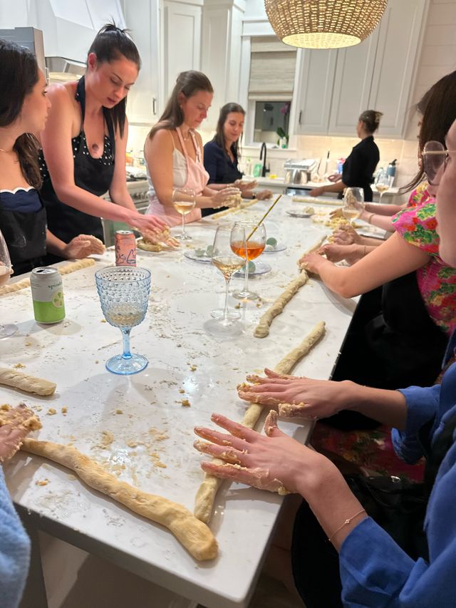 Friends' baking party in a modern white kitchen: people rolling long bread dough ropes on a marble island with floury hands, wine glasses and drinks under a woven pendant light.