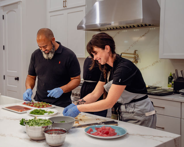 Two people in a modern white kitchen preparing sushi-grade tuna and jalapeño-topped bites on a marble island, smiling and wearing aprons and blue gloves