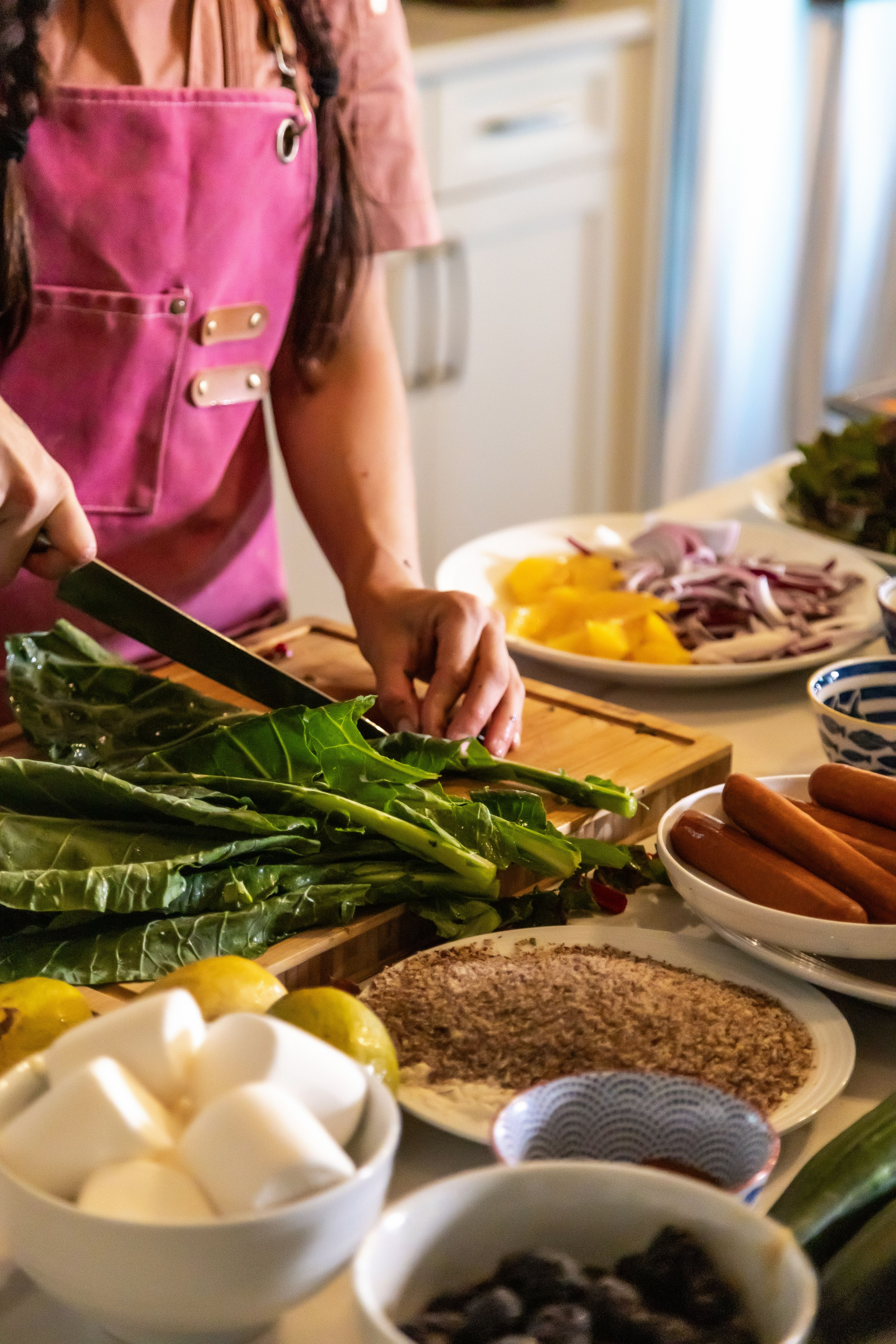 Meal prep in a home kitchen: hands in a pink apron chopping leafy greens on a wooden cutting board surrounded by bowls of eggs, lemons, sausages, sliced peppers, onions and spices.