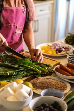 Meal prep in a home kitchen: hands in a pink apron chopping leafy greens on a wooden cutting board surrounded by bowls of eggs, lemons, sausages, sliced peppers, onions and spices.