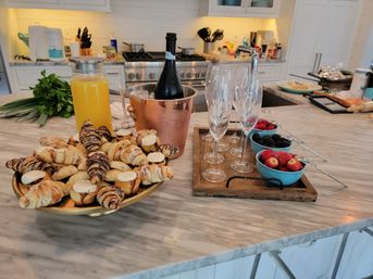 Brunch spread on a marble kitchen island with a platter of croissants and pastries, pitcher of orange juice, chilled champagne in a copper bucket, row of champagne flutes and bowls of strawberries, blackberries and raspberries.