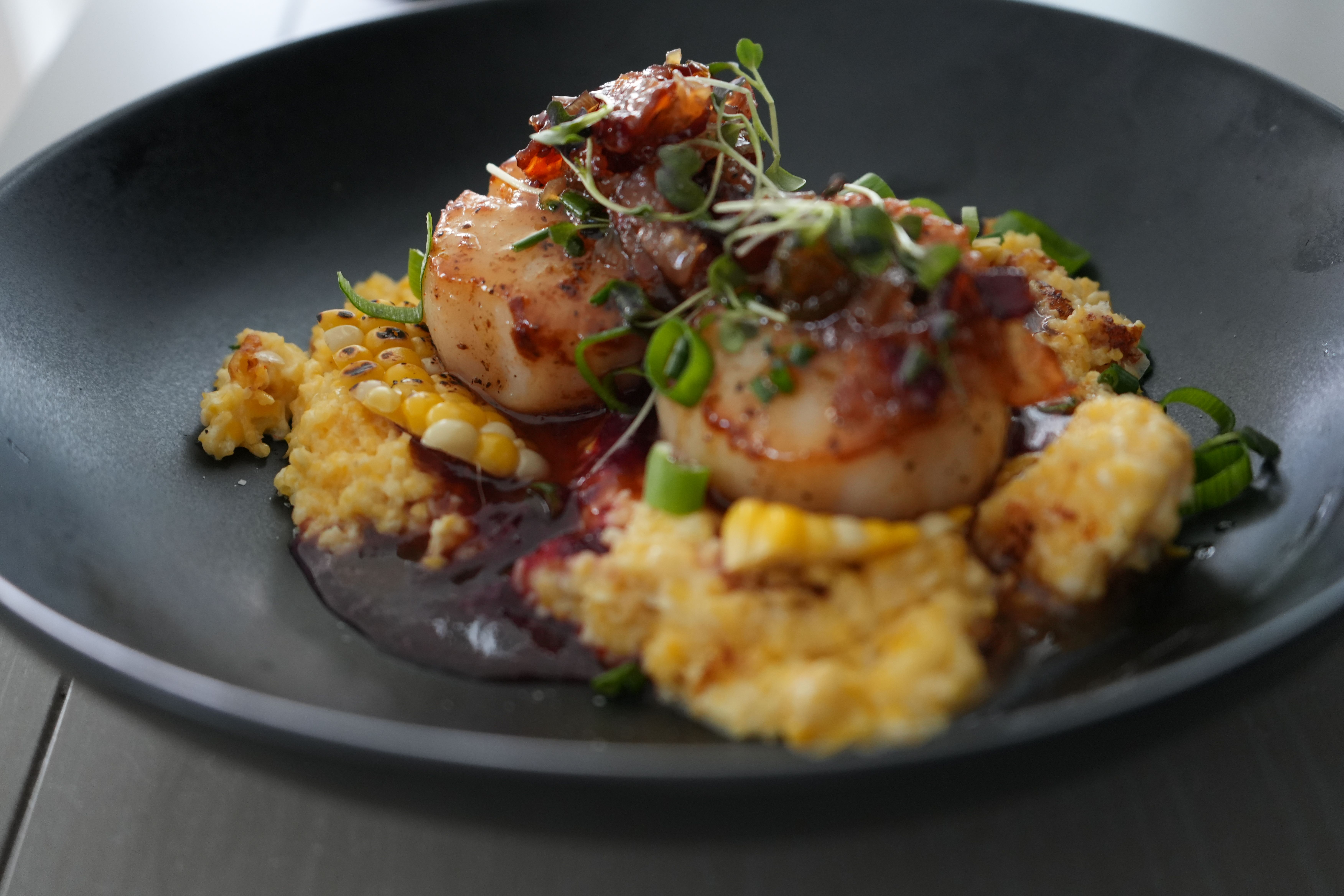 Two seared scallops on creamy corn polenta with charred corn kernels, rich red glaze, sliced green onions and microgreens on a matte black plate
