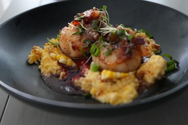 Two seared scallops on creamy corn polenta with charred corn kernels, rich red glaze, sliced green onions and microgreens on a matte black plate