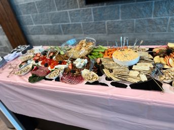 Indoor grazing table at an event with crackers and cheese dip, charcuterie, pistachios, fresh fruit, carrots and celery on a pink tablecloth against a gray stone wall