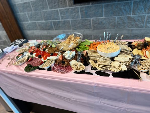 Indoor grazing table at an event with crackers and cheese dip, charcuterie, pistachios, fresh fruit, carrots and celery on a pink tablecloth against a gray stone wall