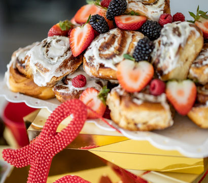 Iced cinnamon rolls piled on a white cake stand, topped with fresh strawberries, blackberries and raspberries, styled on a mirrored gold surface with a red 'love' decoration — brunch dessert display.