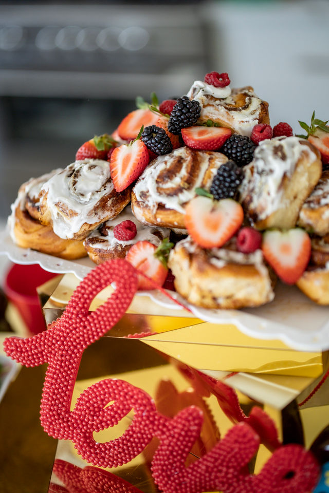 Iced cinnamon rolls piled on a white cake stand, topped with fresh strawberries, blackberries and raspberries, styled on a mirrored gold surface with a red 'love' decoration — brunch dessert display.