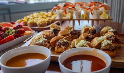 Mouthwatering catered buffet on a wooden table: mini pulled-pork sliders with coleslaw, shrimp cocktail cups with lemon wedges, assorted hors d'oeuvres, fresh fruit platter and two bowls of soup