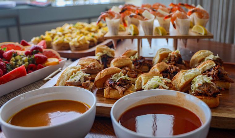 Mouthwatering catered buffet on a wooden table: mini pulled-pork sliders with coleslaw, shrimp cocktail cups with lemon wedges, assorted hors d'oeuvres, fresh fruit platter and two bowls of soup