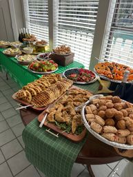 Sunlit indoor brunch buffet on a green-check tablecloth with waffles, fried chicken tenders, assorted pastries, watermelon and berry platters, sliced oranges and bottles of sparkling wine.