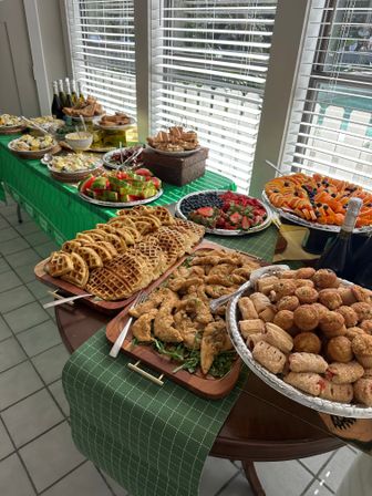 Sunlit indoor brunch buffet on a green-check tablecloth with waffles, fried chicken tenders, assorted pastries, watermelon and berry platters, sliced oranges and bottles of sparkling wine.
