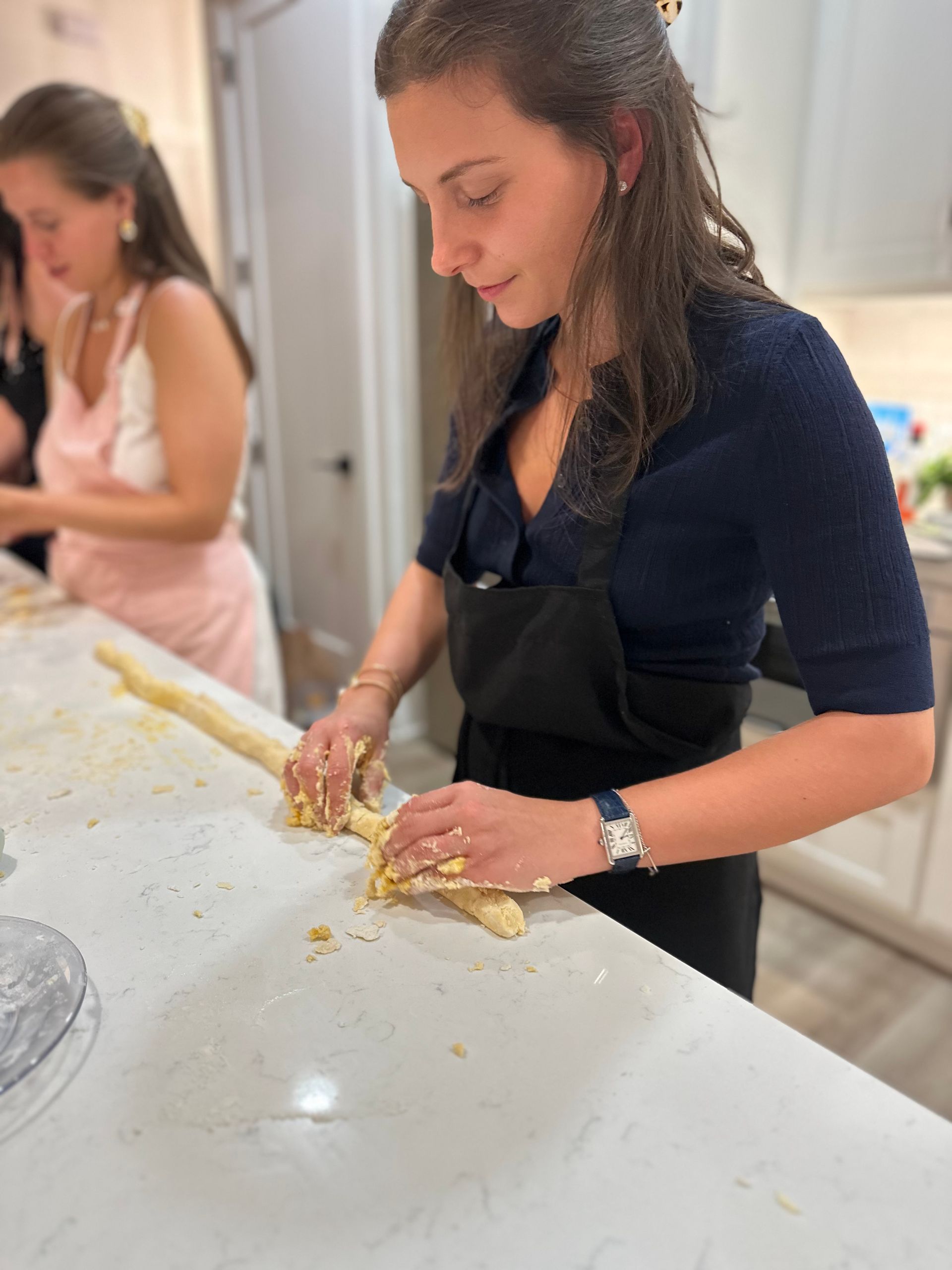 Woman in a black apron rolling dough into a long rope on a marble kitchen island during a home baking session, hands dusted with flour