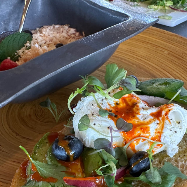 Close-up gourmet brunch: poached egg drizzled with chili oil on avocado toast topped with blueberries and microgreens, served on a wooden plate with a metal bowl of granola nearby