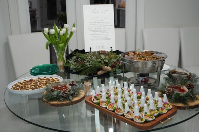 Festive catered buffet on a round glass table with white tulips, pine-and-wood centerpieces, rows of appetizer spoons, a seafood chafing dish and bite-size pastries