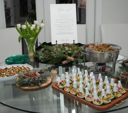 Festive catered buffet on a round glass table with white tulips, pine-and-wood centerpieces, rows of appetizer spoons, a seafood chafing dish and bite-size pastries