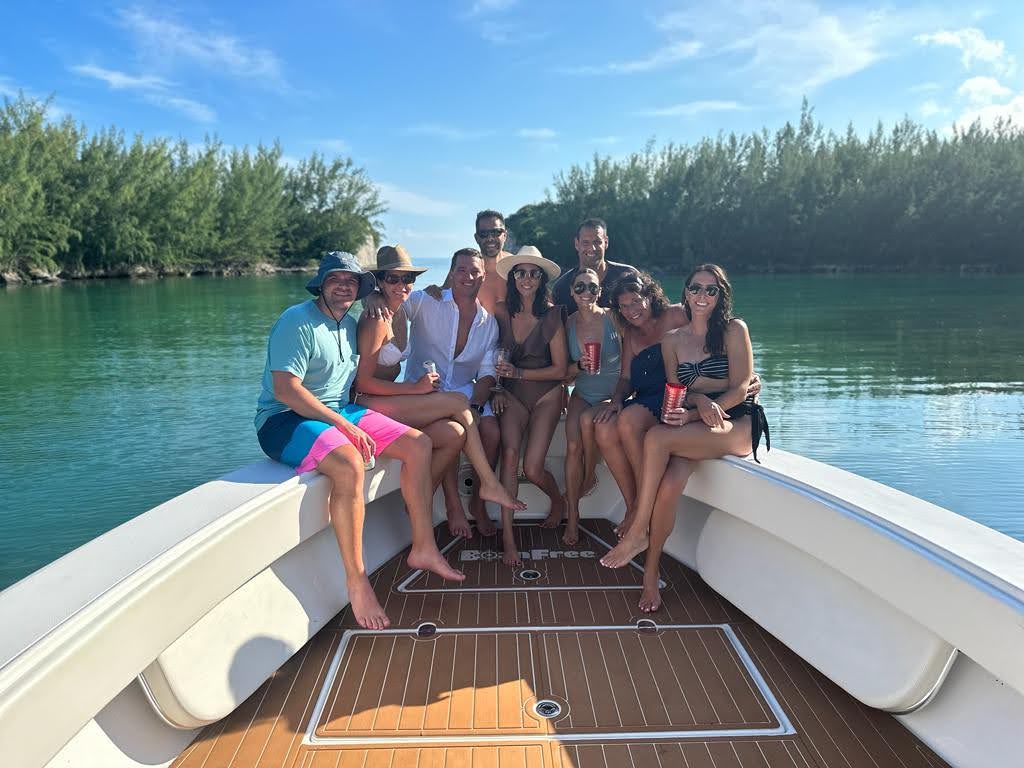 Smiling group of friends in swimsuits sitting on the bow of a motorboat, holding drinks under a bright blue sky on calm turquoise water with a tree-lined shoreline.