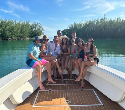 Smiling group of friends in swimsuits sitting on the bow of a motorboat, holding drinks under a bright blue sky on calm turquoise water with a tree-lined shoreline.