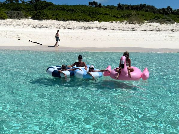 Group on inflatable floats — a pink flamingo and blue-and-white tubes — drifting in crystal-clear turquoise water off a white-sand tropical beach.