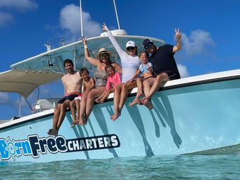Group of adults and children in swimsuits sitting on the side of a light-blue charter boat with legs dangling over clear turquoise water, waving and smiling under a sunny blue sky.