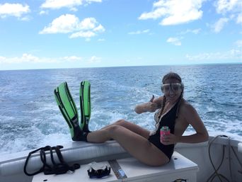 Person in a swimsuit and snorkel mask relaxing on a boat with neon green fins, giving a thumbs-up and holding a canned drink against the open ocean and sunny blue sky.