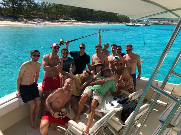 Group of friends on a boat in bright turquoise tropical water near a white-sand beach, holding drinks and a fishing rod on a sunny day.