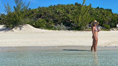 Person in a sun hat and bikini holding a drink stands in clear shallow turquoise water at a white‑sand tropical beach with coastal greenery and a bright blue sky