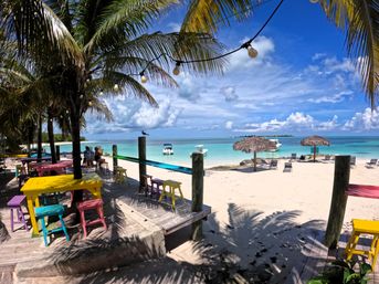 Colorful beachfront bar with painted tables and stools under palm trees and string lights, white-sand tropical island beach with turquoise water, small boats and thatched umbrellas.