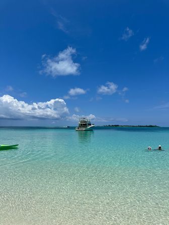 Crystal-clear turquoise lagoon and white-sand beach with an anchored motorboat, a green kayak at the shore, and two swimmers under a bright blue sky with fluffy clouds.